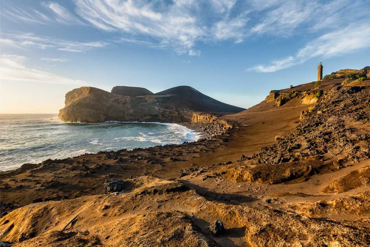 Coastal landscape with rocky sandy beach, ocean waves, cliffs, and a lighthouse on a hill under a partly cloudy sky