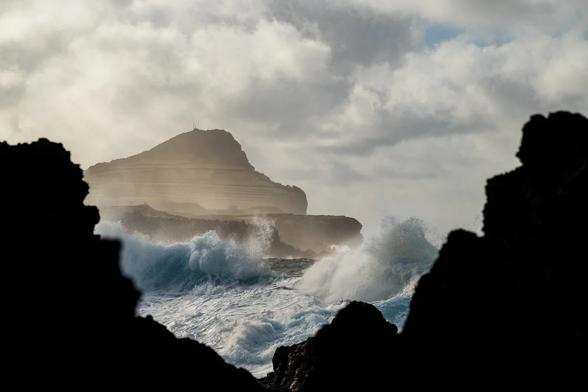 Ocean waves crashing against dark rocky shoreline with a mountain in the background and cloudy sky above.