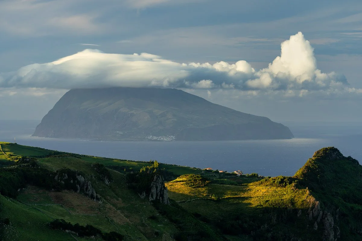 A large island with steep cliffs and a flat top, surrounded by water, with a bank of cloud cover over its summit. In the foreground, green hills with rocky outcroppings and a few scattered buildings are visible, under a partly cloudy sky.