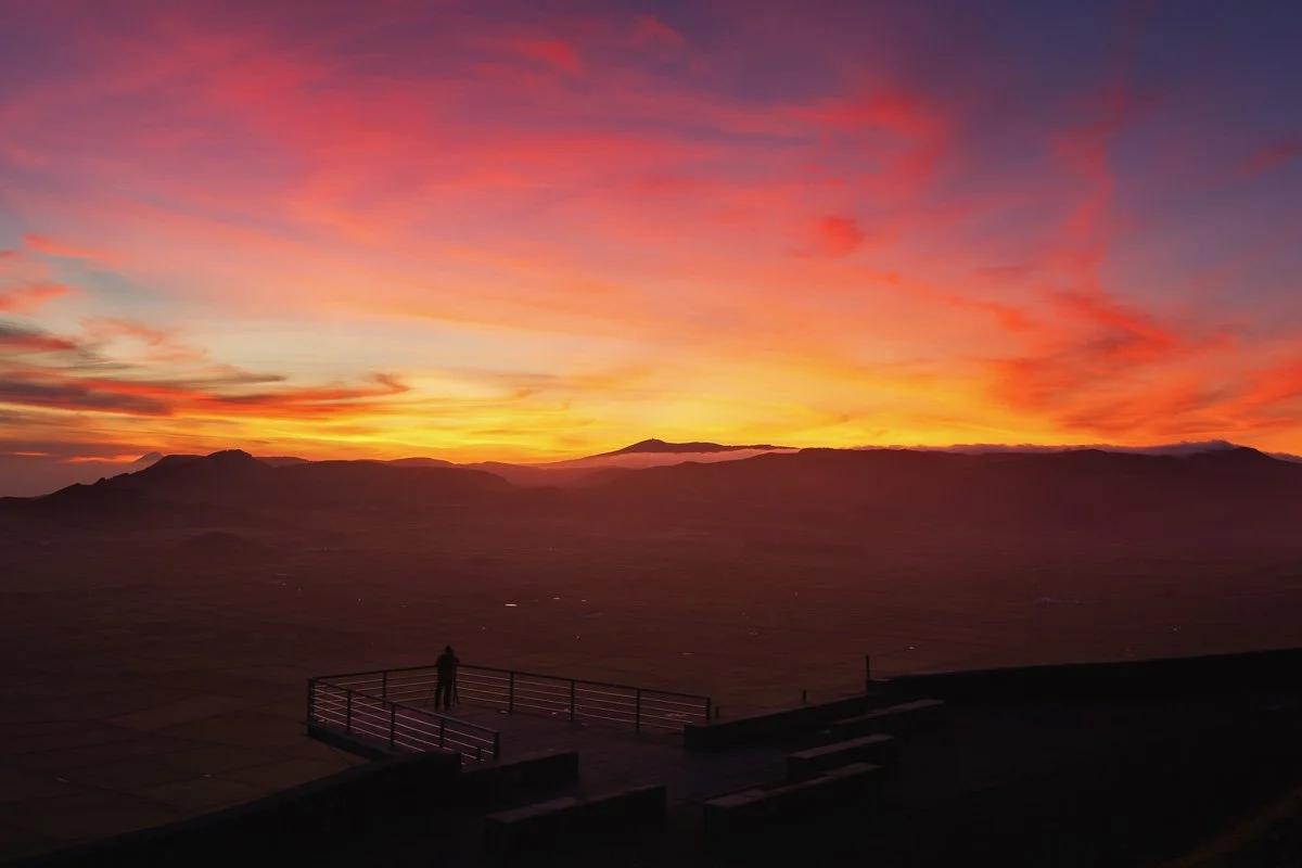 A person standing on an observation deck overlooking a vast landscape at sunset with vibrant orange, pink, and purple clouds.