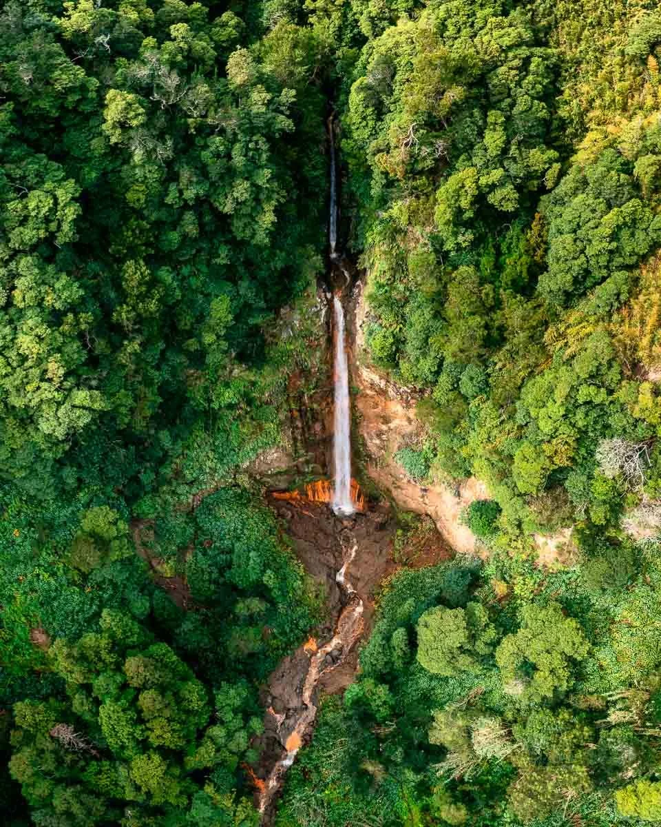 An aerial view of a tall, narrow waterfall in a dense green forest with a rocky stream below.