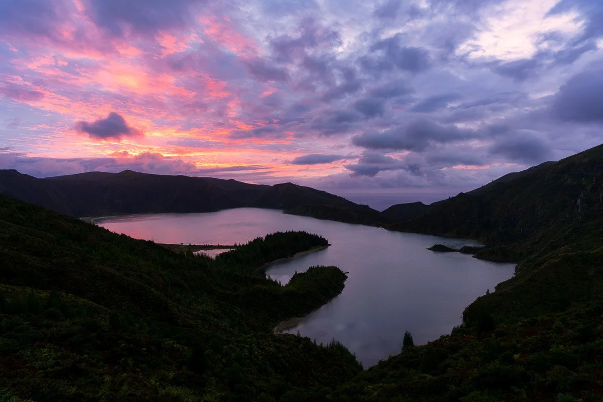 A scenic landscape of a lake surrounded by green mountains under a colorful sunset sky with purple and pink clouds.