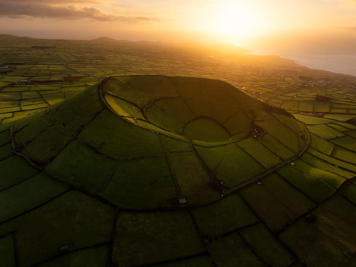 Aerial view of rolling green hills with a large, oval-shaped terraced field at sunset, overlooking a patchwork of fields and the ocean in the distance.