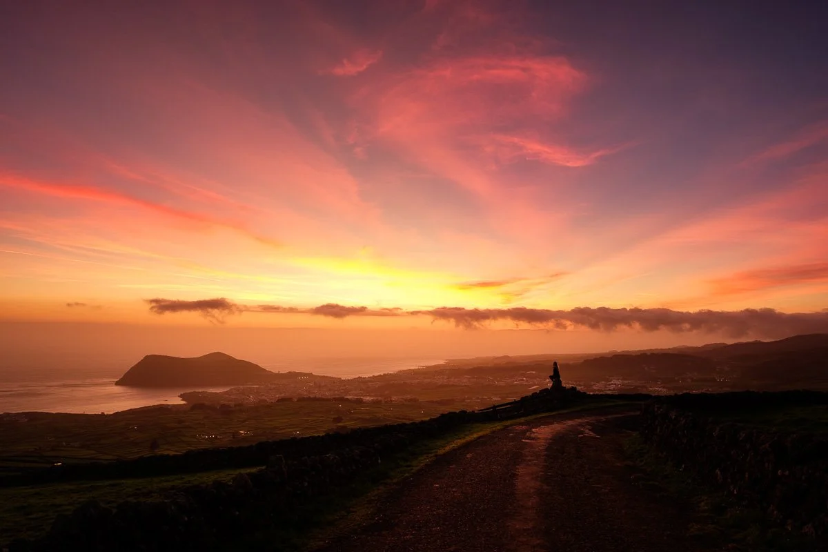 Sunset over a coastal landscape with vibrant pink, orange, and purple sky, a distant mountain or island, and a dirt path leading to a small monument or statue on a hill.