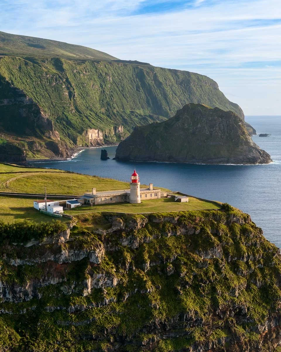 A coastal scene with a lighthouse on a grassy cliff, overlooking the ocean and rugged green cliffs and rock formations in the distance.