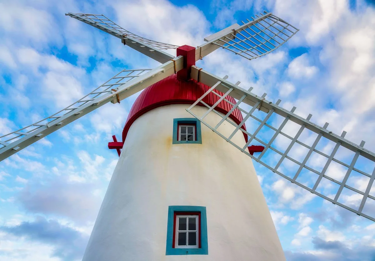 A tall white windmill with red accents and large wooden blades against a cloudy blue sky.