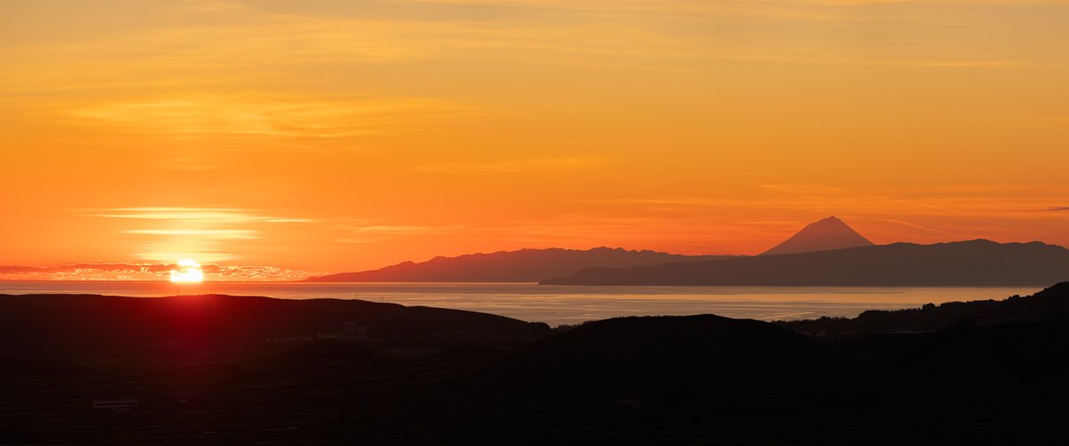 Sunrise over a body of water with distant mountains and a volcano in the background, colorful sky with orange and yellow hues.