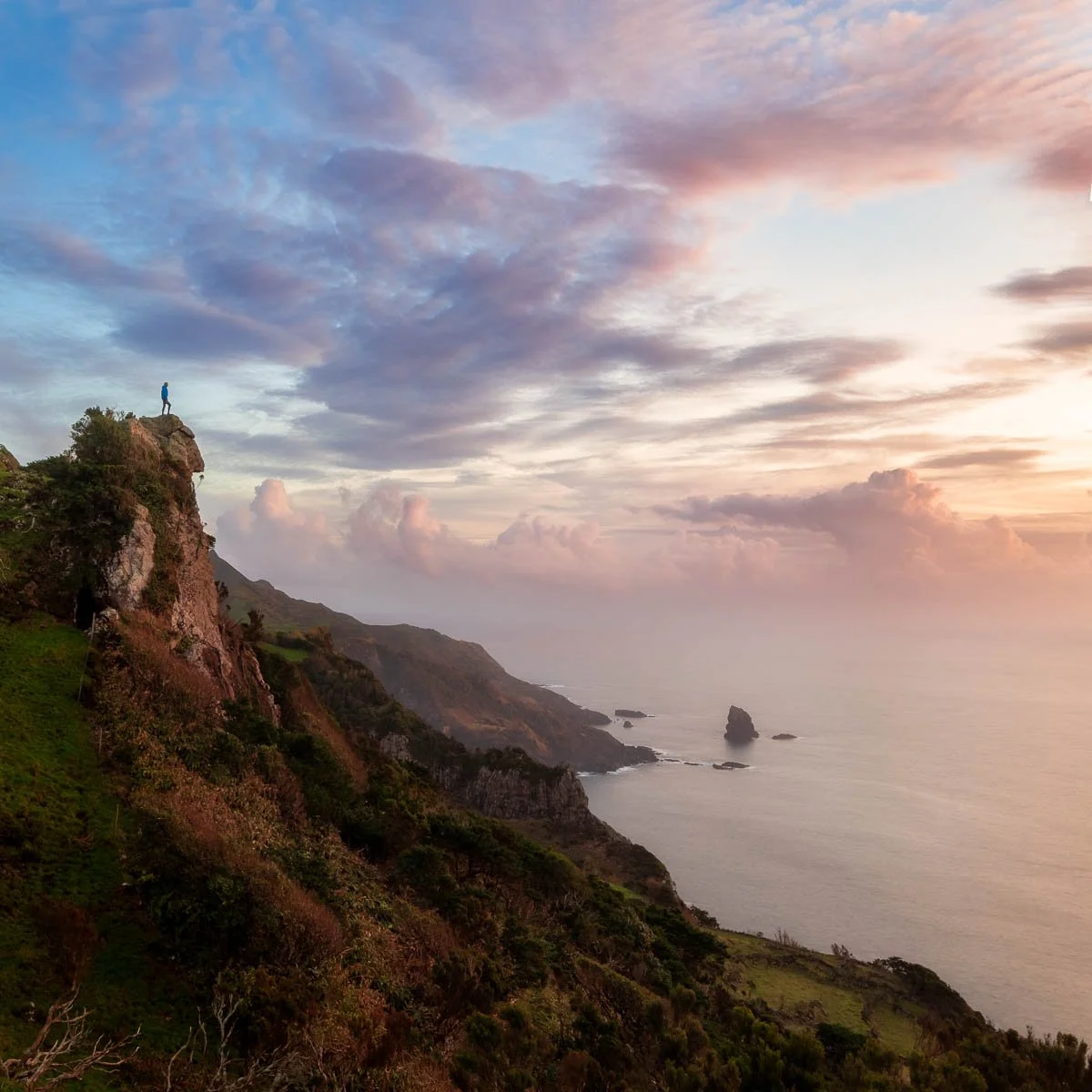 A person standing on a rocky cliff overlooking the ocean at sunset, with clouds and colorful sky in the background.