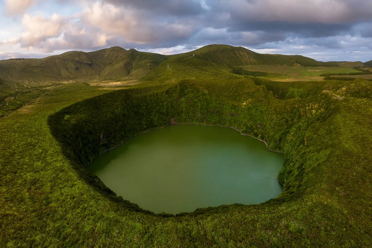 A large, green crater lake surrounded by lush, grassy hills with rolling mountains in the background under a partly cloudy sky.
