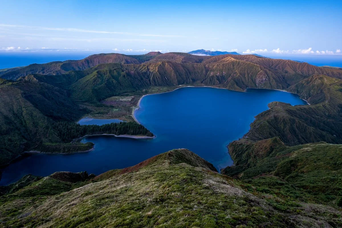 Aerial view of a volcanic crater with a large blue lake inside, surrounded by green and brown mountain ridges under a blue sky.