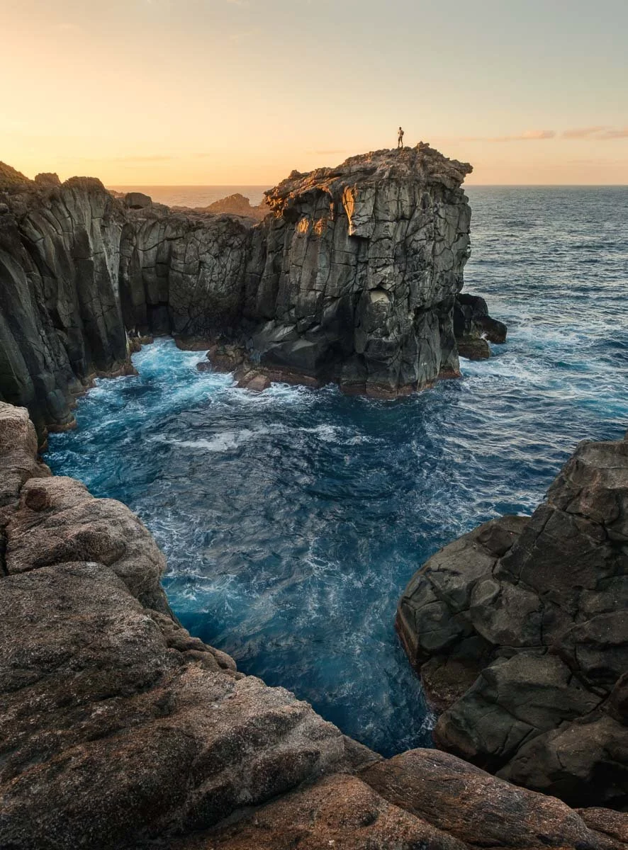 Person standing on top of a large rocky cliff by the ocean at sunset, with waves crashing below and other rocky formations around.