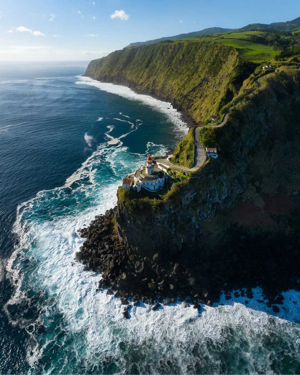 An aerial view of a lighthouse on a rocky cliff overlooking the ocean, with waves crashing below and green hills in the background.