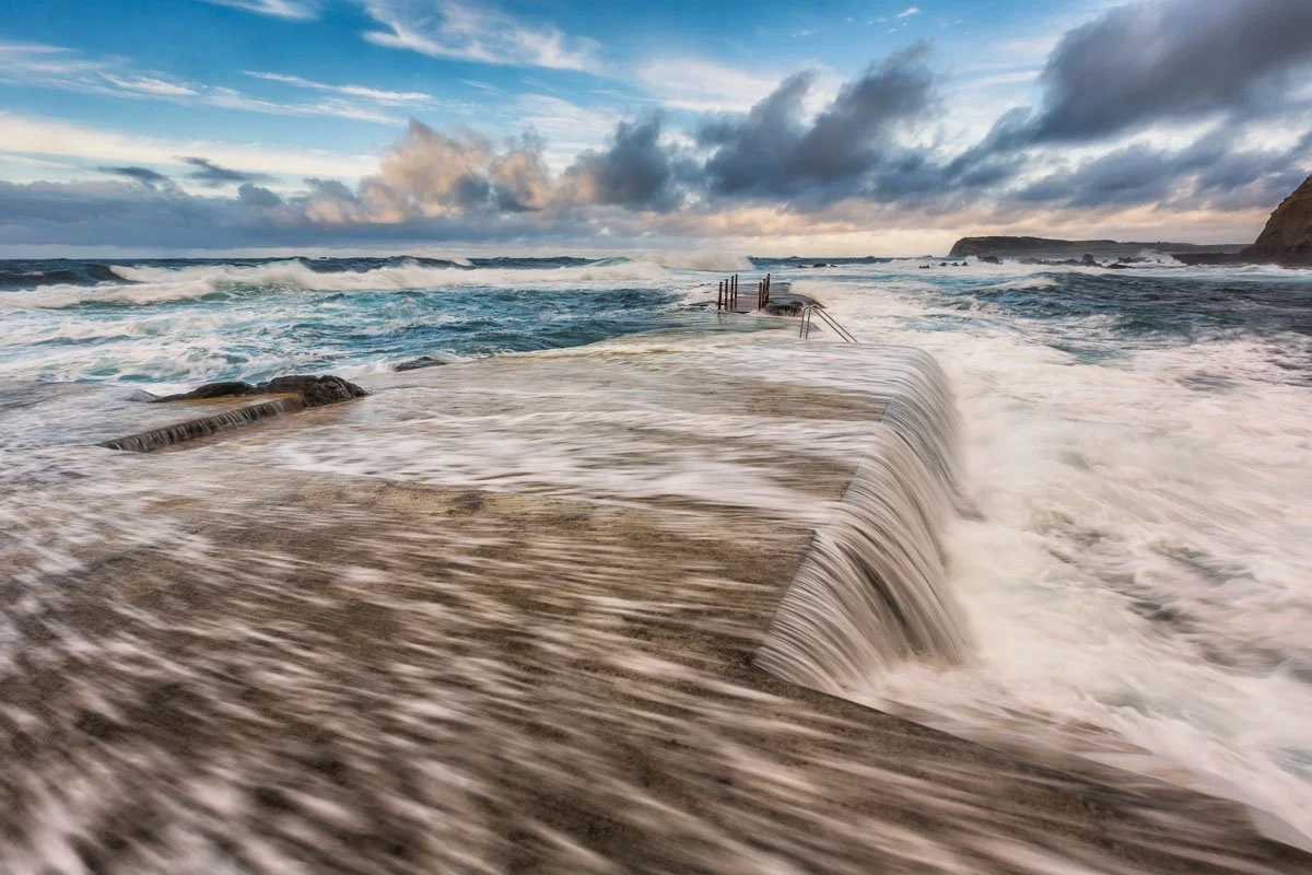 Waves crashing over a concrete pier at the beach with a cloudy sky.