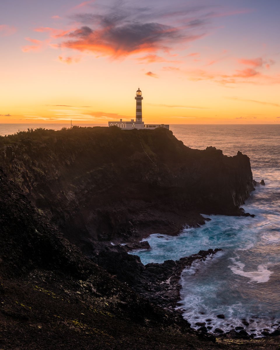 A lighthouse on a cliff during sunset with a colorful sky and ocean waves below.
