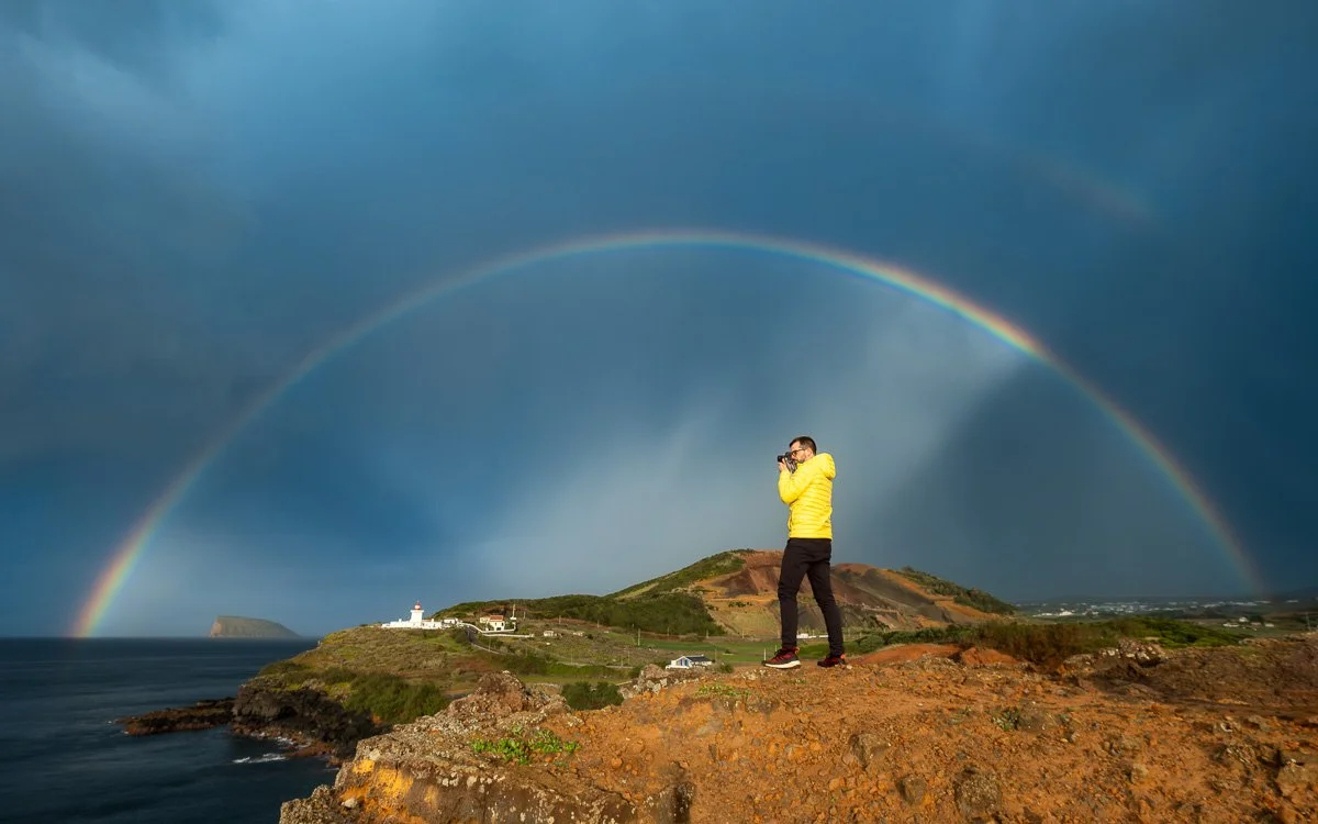 Person in yellow jacket taking a photograph on a rocky hillside with a rainbow over the ocean and coastline in the background.