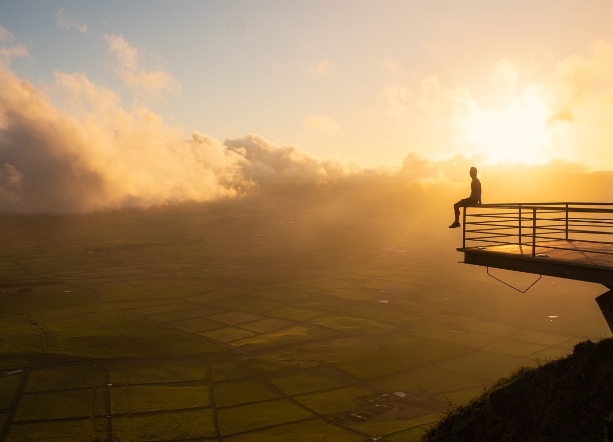 A person sitting on the edge of a rooftop overlooking a landscape with clouds and a sunset in the distance.