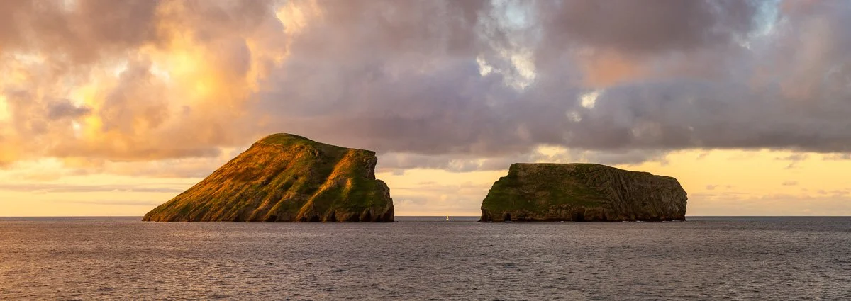 Two large, grass-covered sea stacks in the ocean during sunset with cloudy sky.