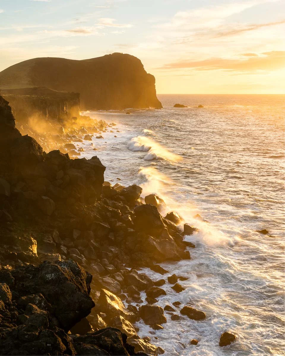 Sunset over rocky coastal cliffs with waves crashing on the shore.