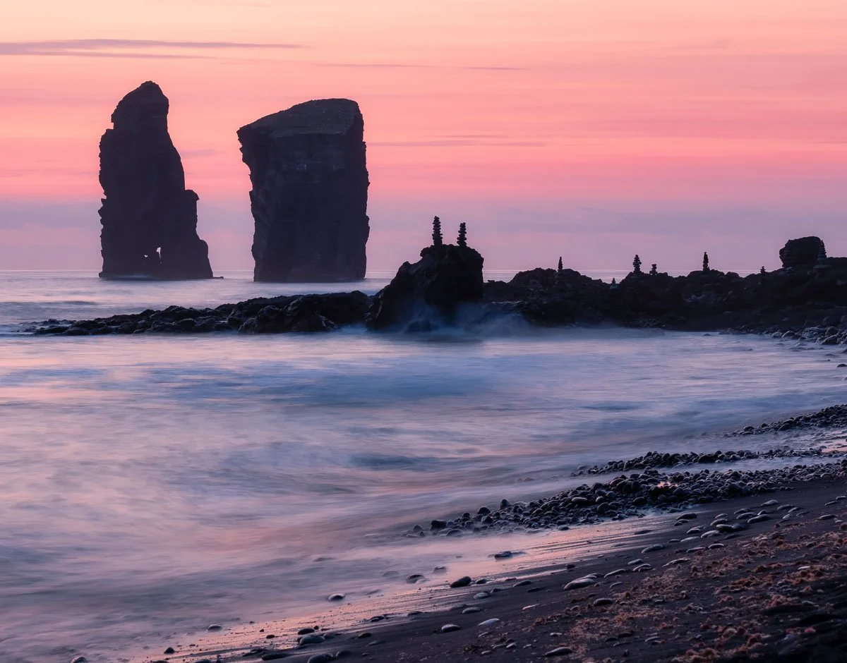 Sunset at the beach with two large sea stacks and smaller formations, gentle waves, and a rocky surf.