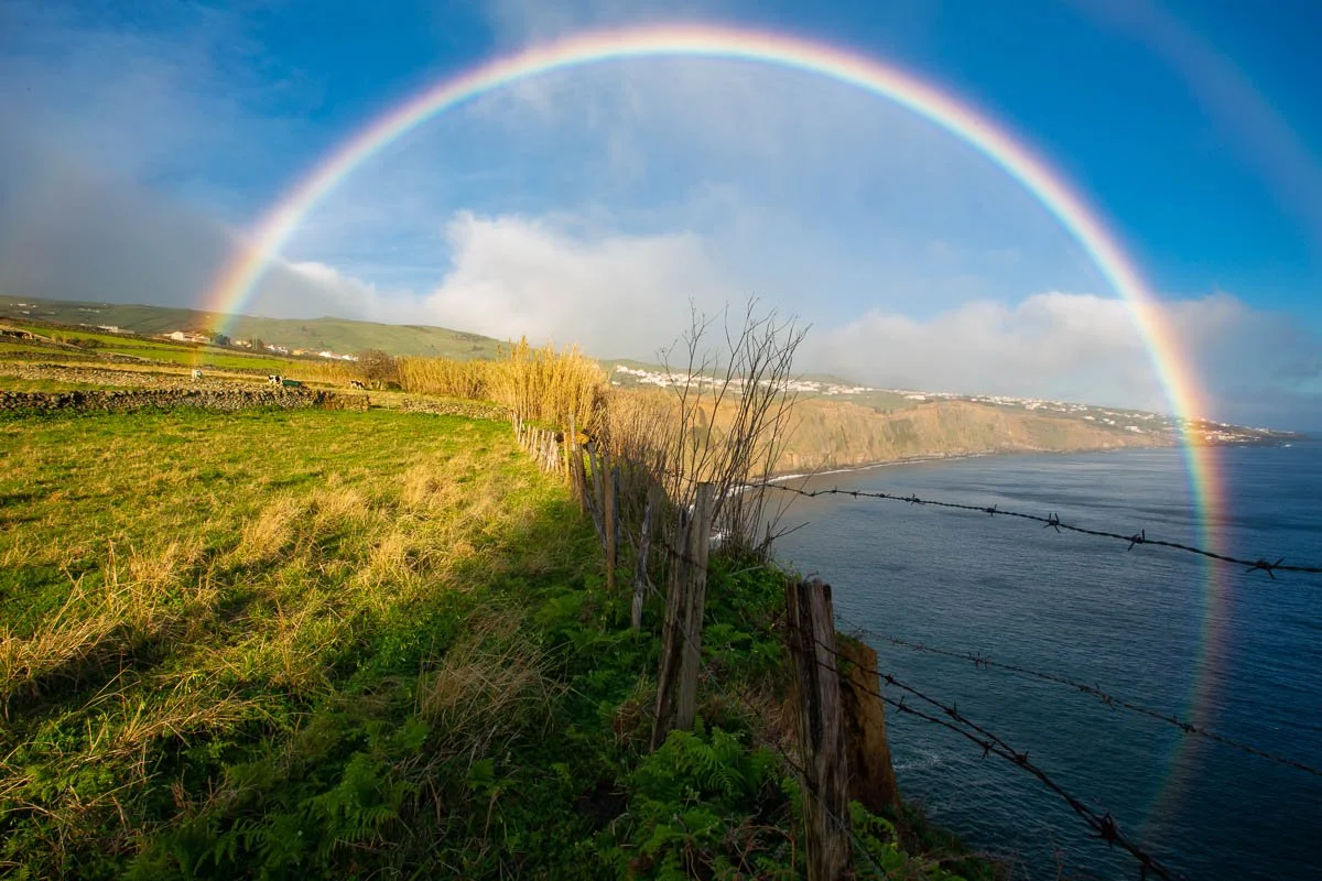 A vibrant rainbow arches over a coastal landscape, with green fields and a barbed wire fence along a cliff near the ocean under a partly cloudy sky.
