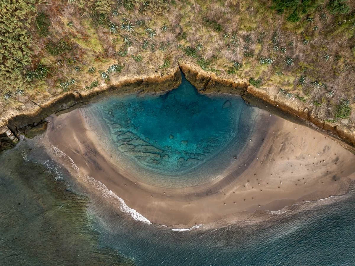 Aerial view of a coastal landscape featuring a natural sea lagoon with blue water, sandy beach, rocky shoreline, and lush greenery with trees and shrubs.