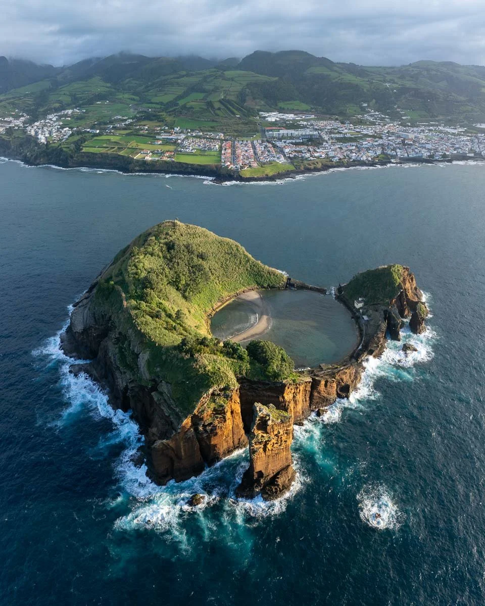 Aerial view of a rocky island with a lagoon, surrounded by ocean, with a coastal town and green hills in the background.
