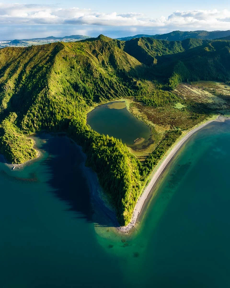 Aerial view of lush green mountains with a body of water in a cove and a long strip of road along the shoreline.