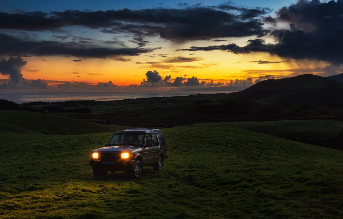 A vehicle with its headlights on parked on a grassy hill during sunset, with rolling hills, mountains, and cloudy sky in the background.