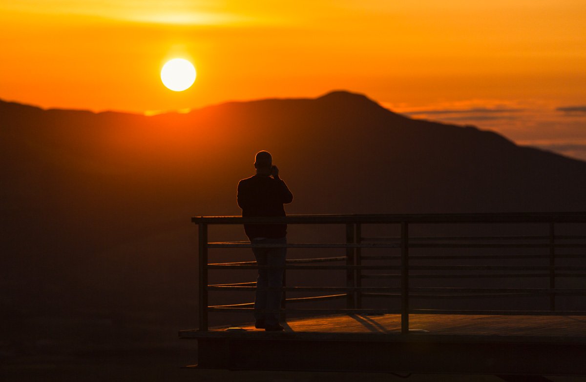 Person standing on a viewing platform, talking on a phone at sunset with mountains in the background.