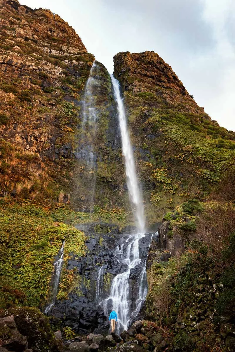 Tall waterfall cascading down a rocky mountain surrounded by green and yellow foliage, with a person standing at the bottom in a blue jacket.