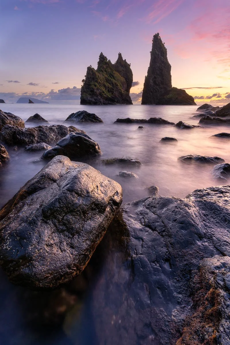 Rocky shoreline at sunset with two large sea stacks in the distance and a colorful sky.