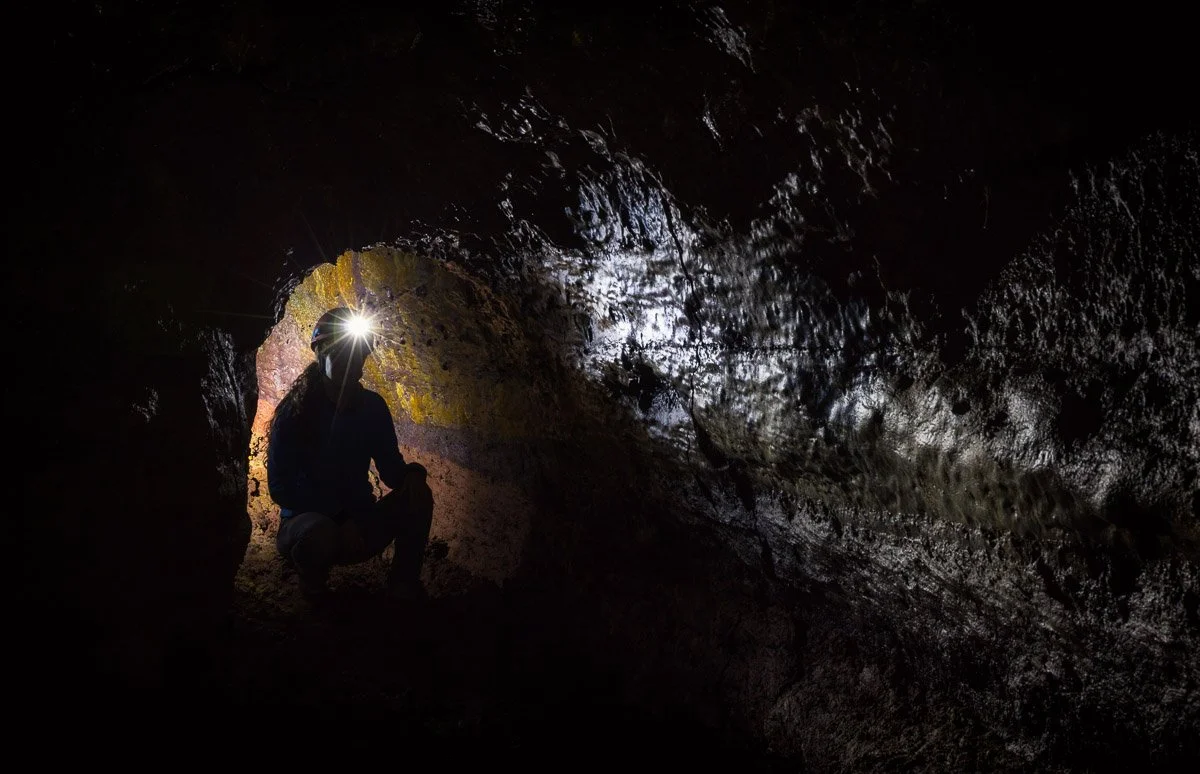 A person wearing a helmet and crouching inside a dark cave with daylight shining through the entrance.