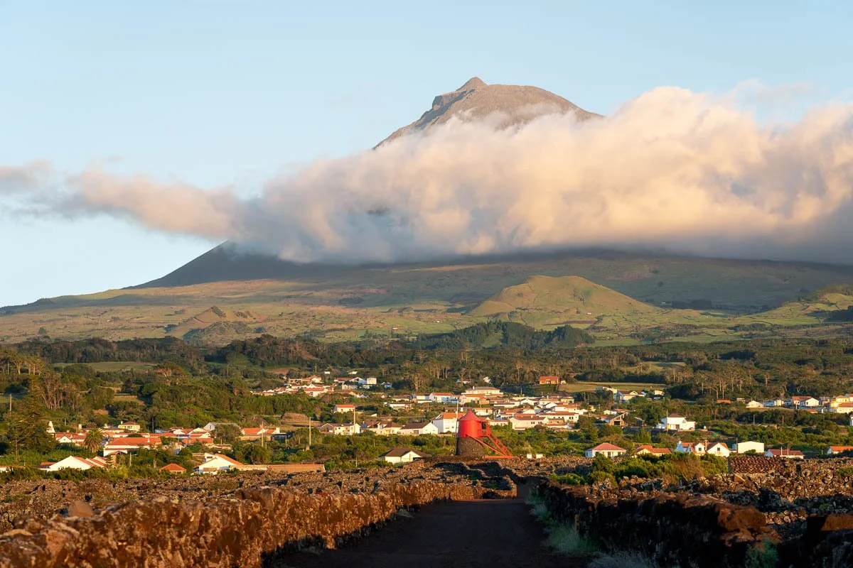 A volcanic mountain with a cloud of smoke around its middle, overlooking a town with colorful houses and lush green hills.