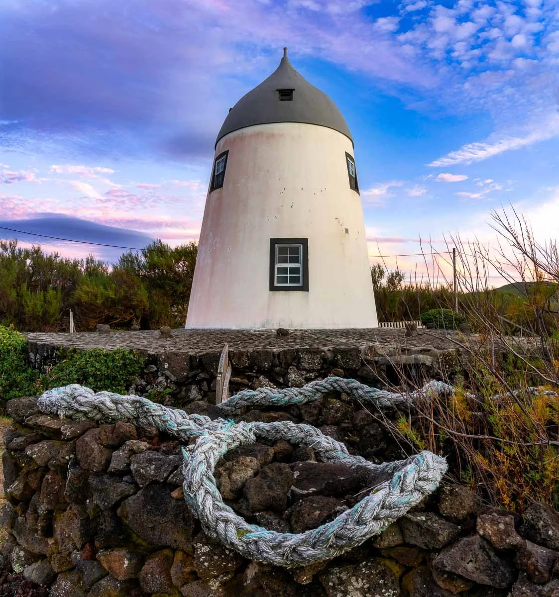 A small white lighthouse with a dark gray roof standing on a rocky base, with a knotted rope in the foreground and a colorful sunset sky with pink, purple, and blue clouds in the background.