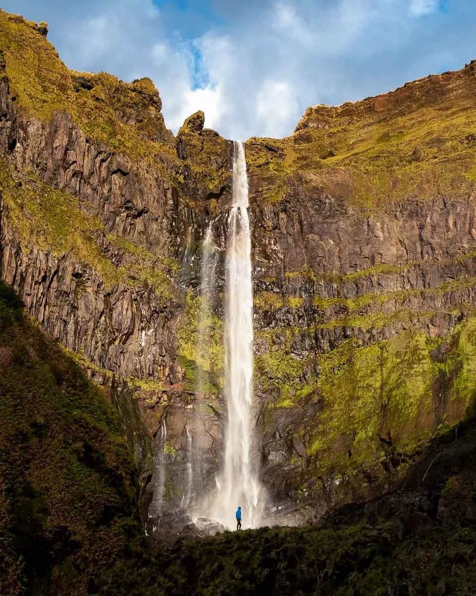 A tall waterfall cascading down a rocky cliffside with patches of green moss and vegetation, a person standing at the base, and a partly cloudy blue sky above.