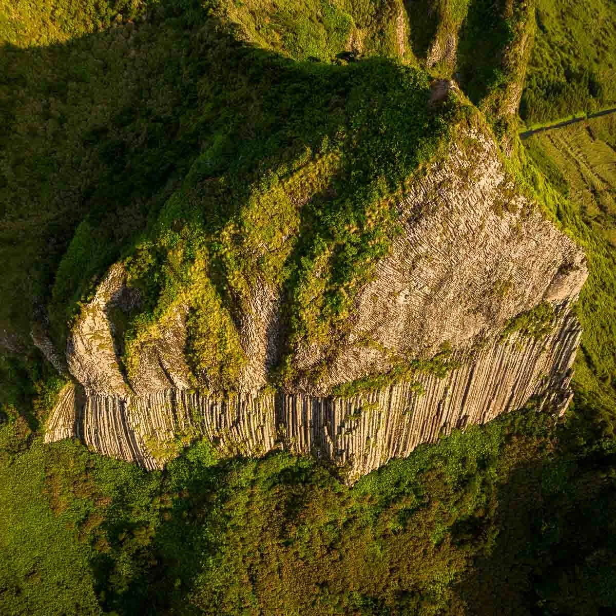 An aerial view of a steep cliff with layers of rock and green vegetation on top.