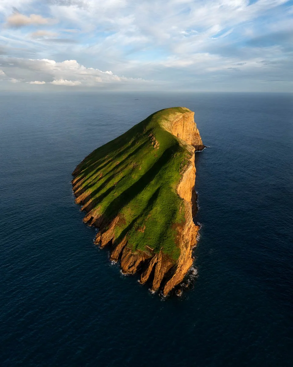 An aerial view of a green, grassy island with steep cliffs surrounded by the ocean, under a partly cloudy sky.