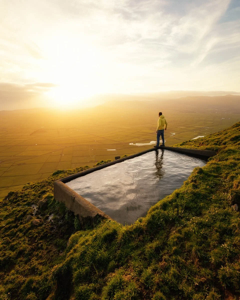 A person standing on the edge of an infinity pool on a grassy hill during sunset, overlooking a vast flat landscape.