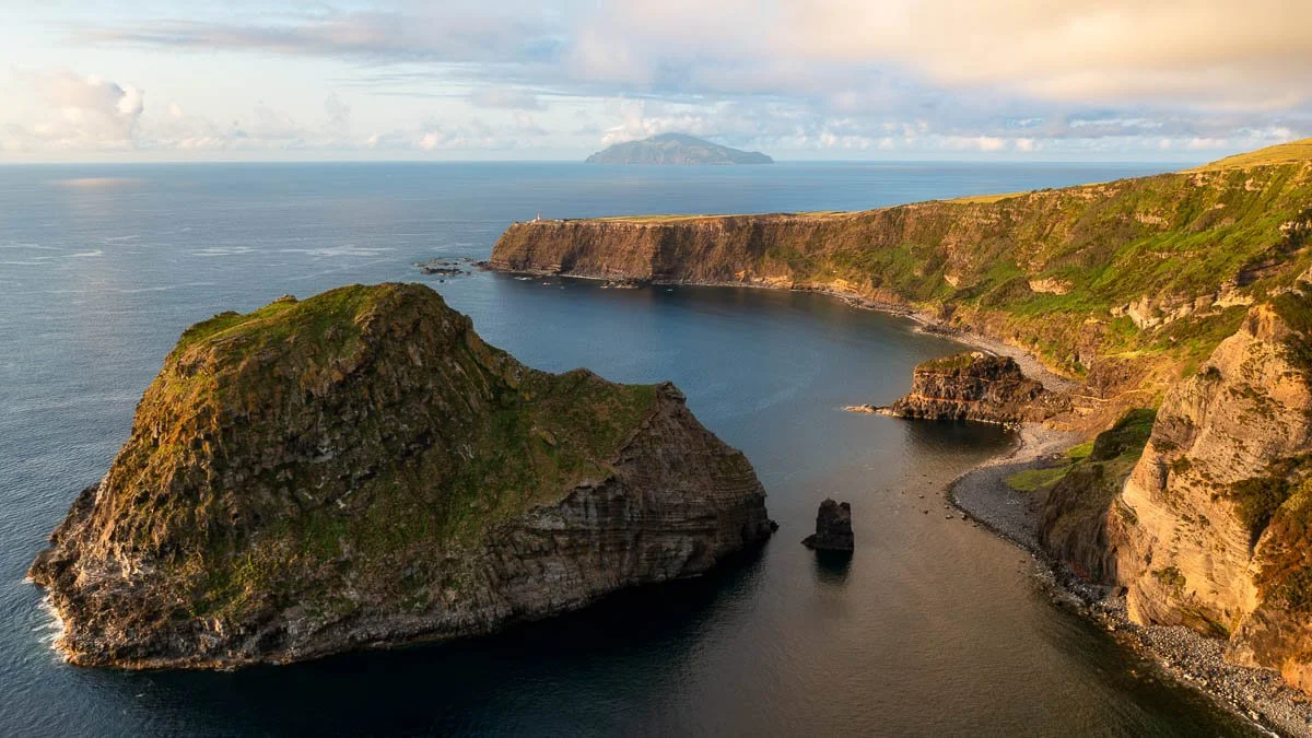 A coastal landscape with rocky cliffs, a cove, and the ocean, with clouds and distant land in the background.
