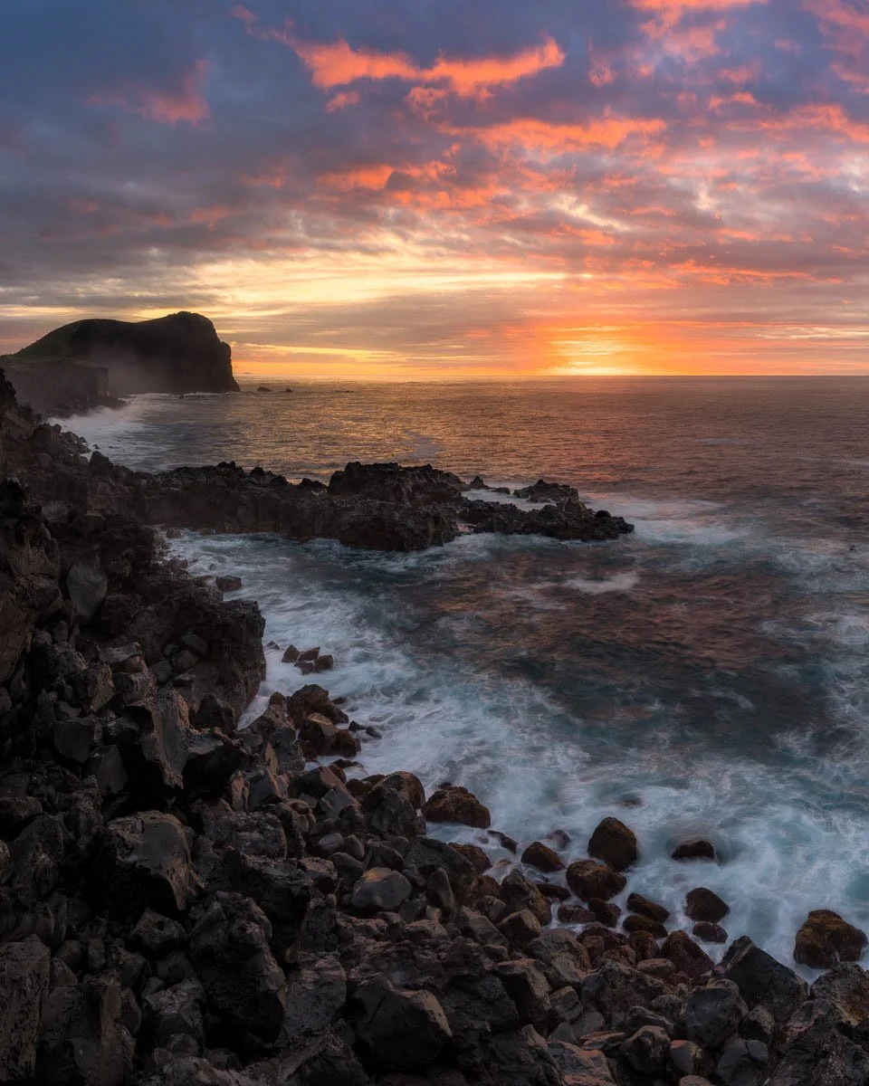 Sunset over the ocean with rocky coastline and colorful sky.