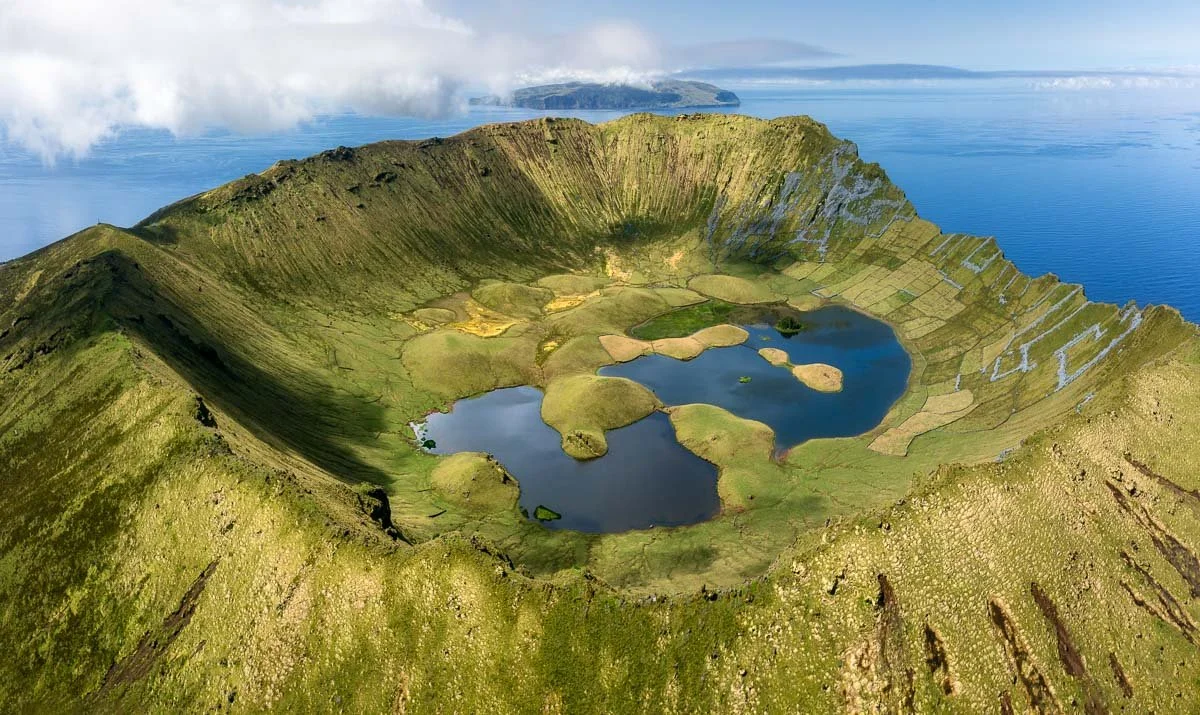 A volcanic crater filled with lakes and green patches, surrounded by grassy slopes and an ocean in the background.