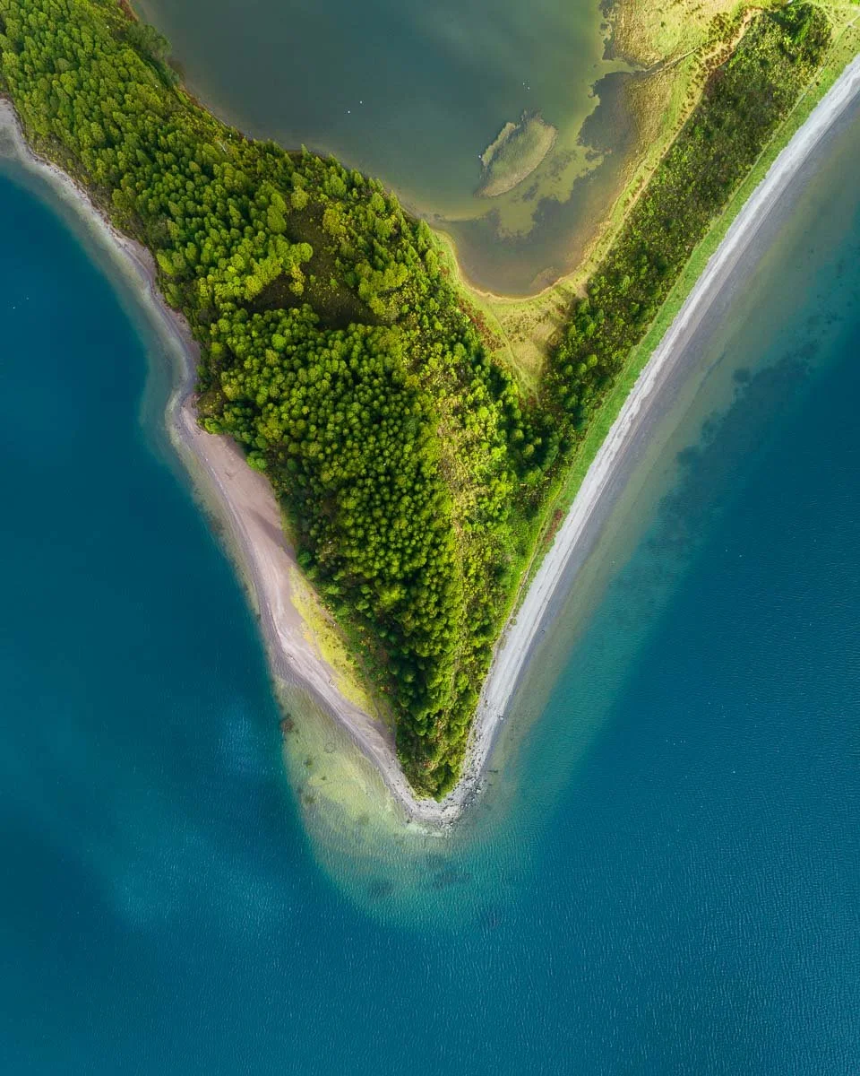 An aerial view of a large, triangular landmass covered in dense green trees, with a narrow strip of shoreline along the edges, surrounded by blue ocean waters. A narrow road runs along one side of the landmass.