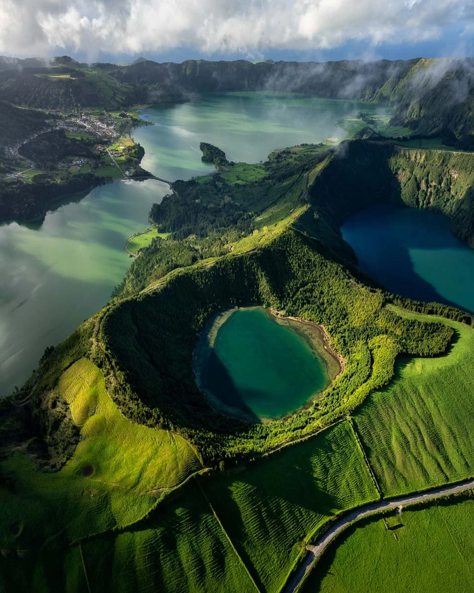 Aerial view of a green volcanic crater surrounded by lush fields and lakes in a mountainous landscape under a cloudy sky.