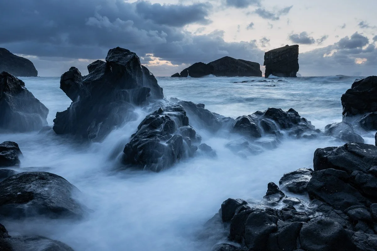 Overcast sky with dark clouds over a rocky coast with large dark rocks in the water and waves crashing around, possibly during dusk or dawn.