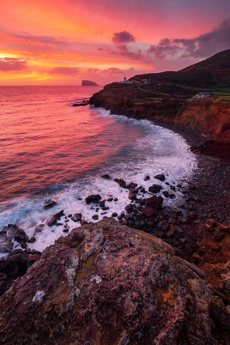 A sunset over the ocean with pink, orange, and purple hues in the sky, waves crashing against a rocky shoreline, and a lighthouse on a distant cliff in the background.