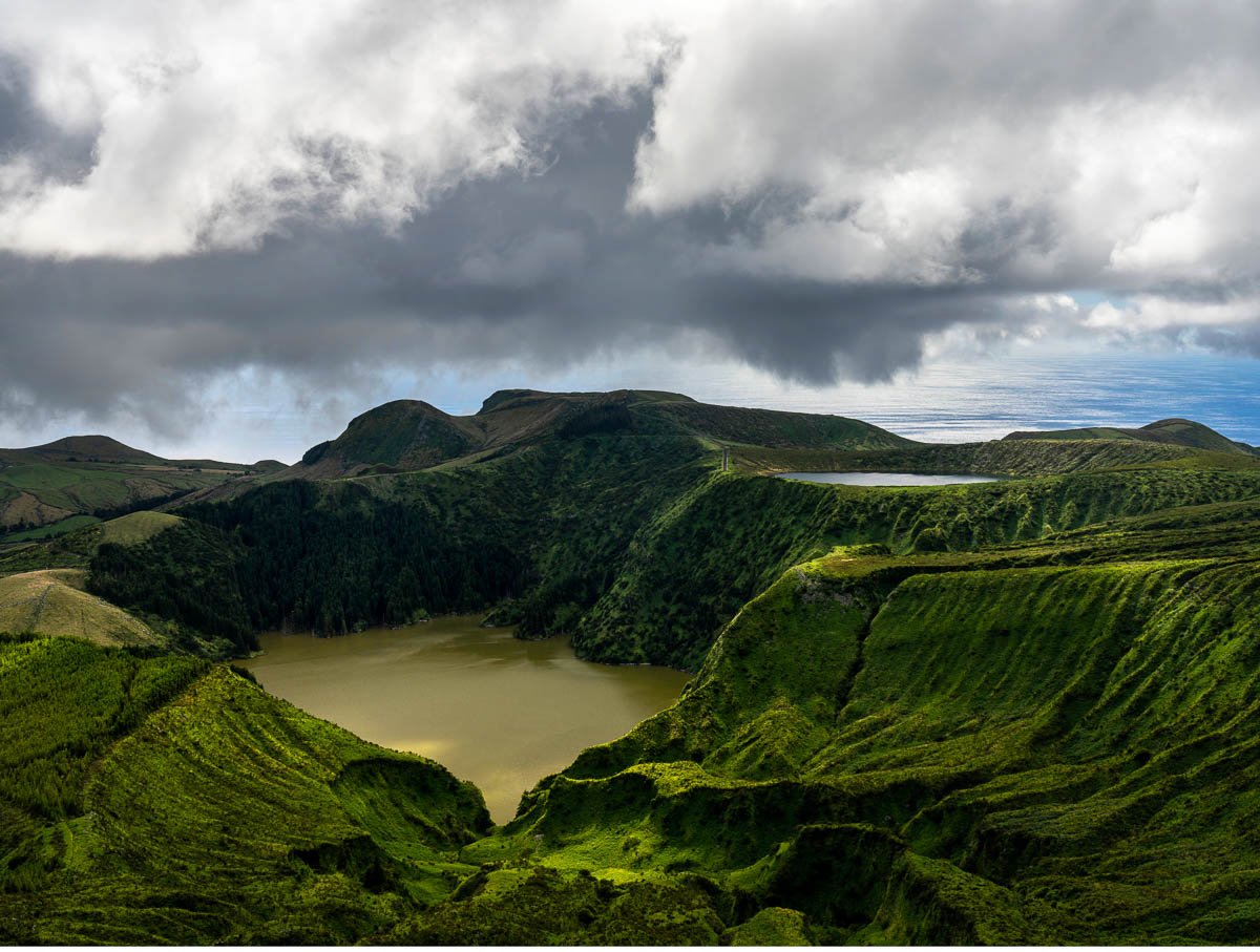 A lush green volcanic crater with two lakes and a mountain in the background under a partly cloudy sky.
