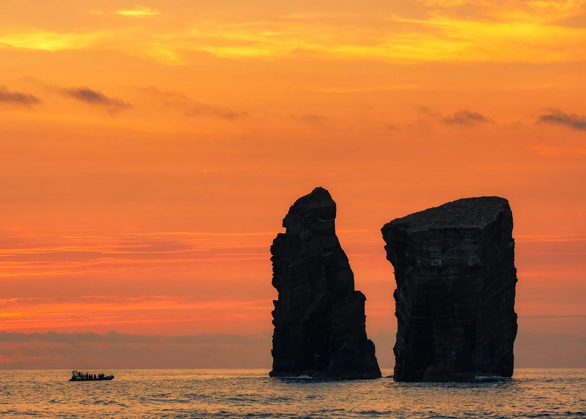 Two large sea stacks rise from the ocean during a colorful sunset sky with orange, yellow, and pink clouds. A small boat with people is seen in the distance.