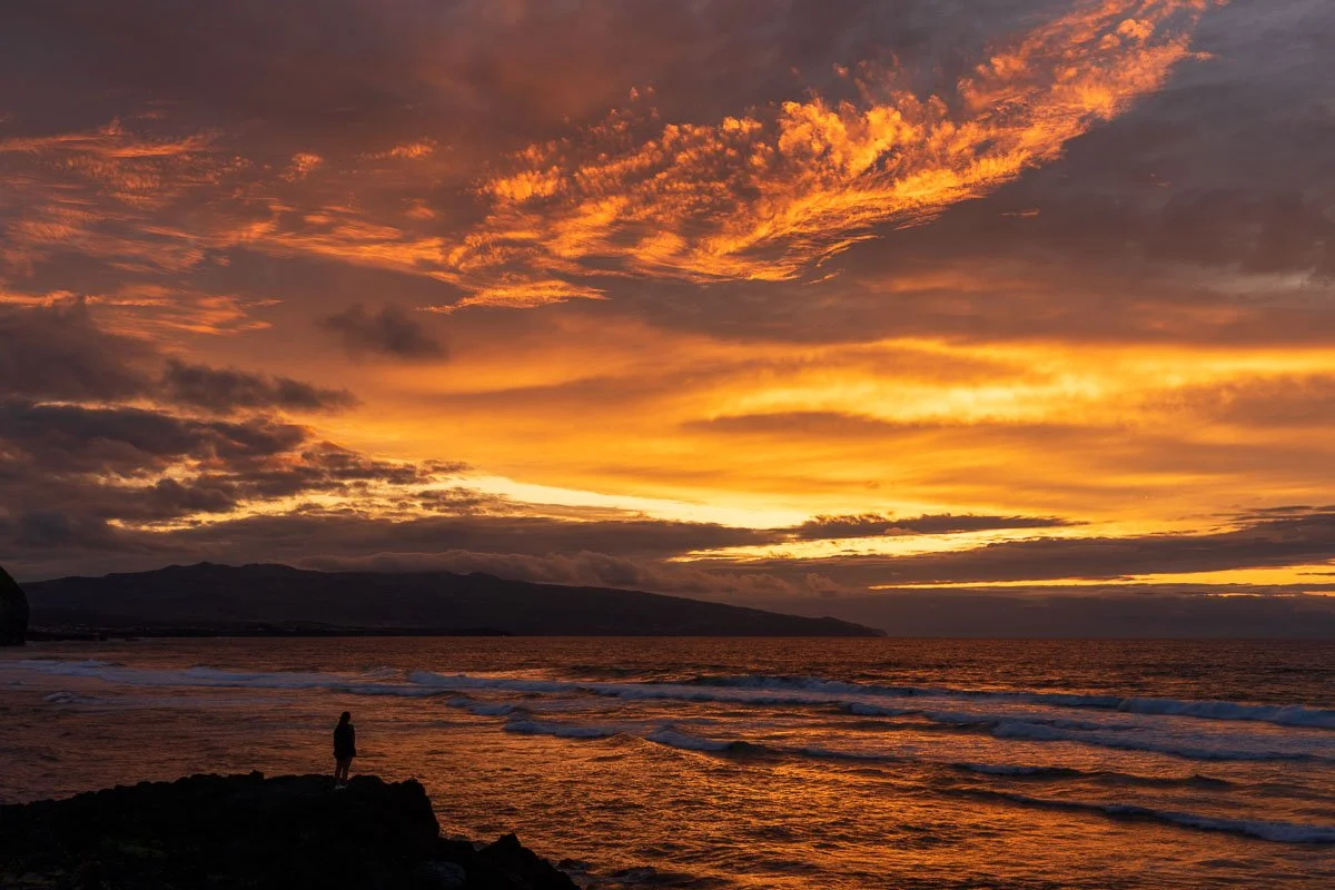 A person standing on rocks by the ocean during a vibrant orange and yellow sunset with clouds in the sky.