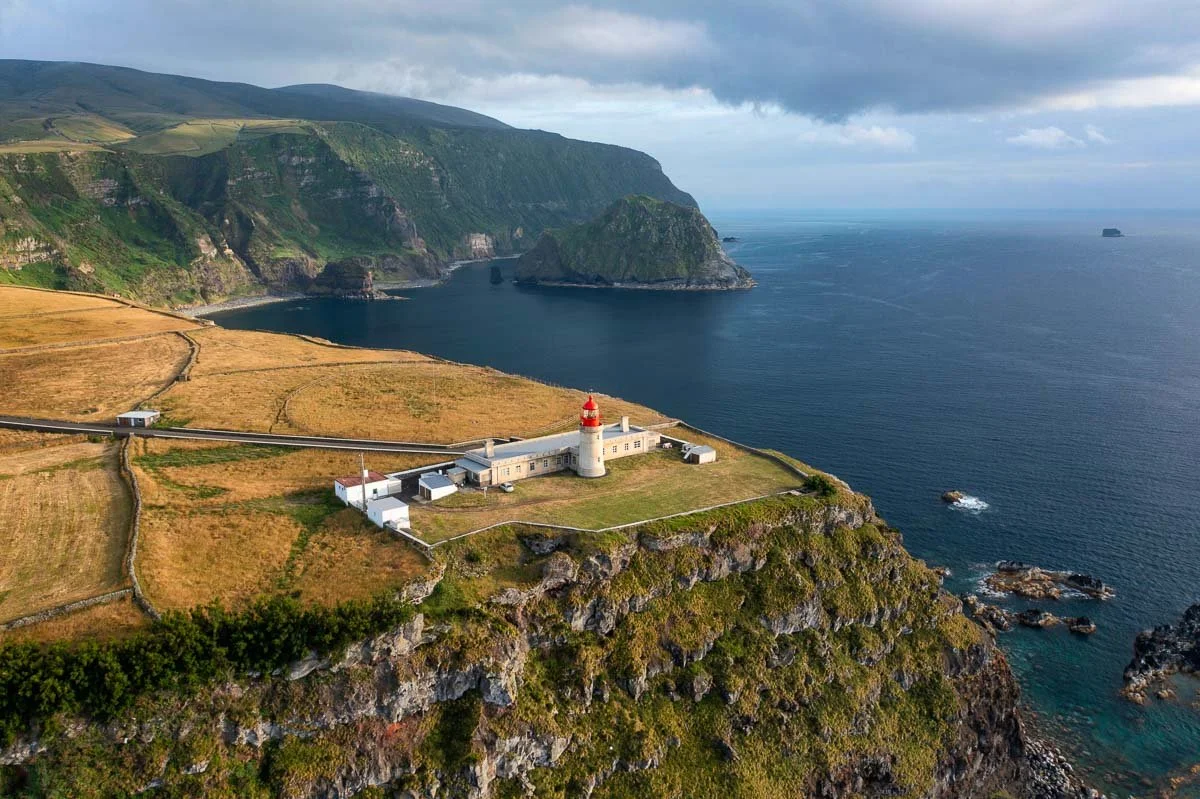 A coastal landscape featuring a lighthouse with a red top, situated on a grassy cliff overlooking deep blue ocean water, with rugged rocks at the base, and green hills and cliffs in the background under a cloudy sky.