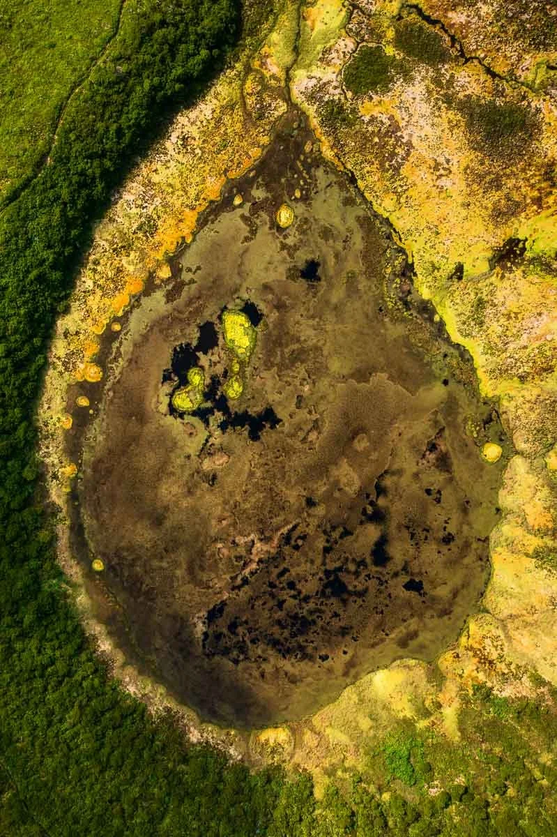 Aerial view of a volcanic crater filled with dark water, surrounded by mossy and lichen-covered rocks and greenery.
